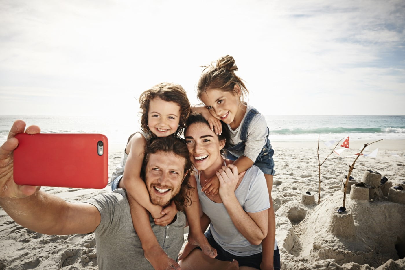 Lykkeleg familie på fire tar ein selfie på stranda, smiler medan dei sit tett saman med eit sandslott i bakgrunnen.