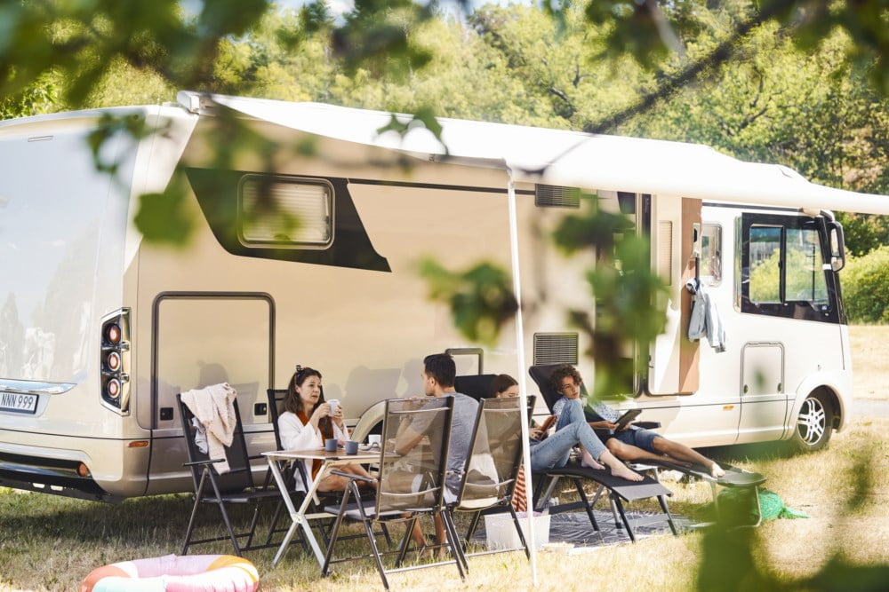 Familie nyter ein campingtur med bubilen parkert i ein solrik skogsgløtt, sit avslappa rundt eit bord under markisa.