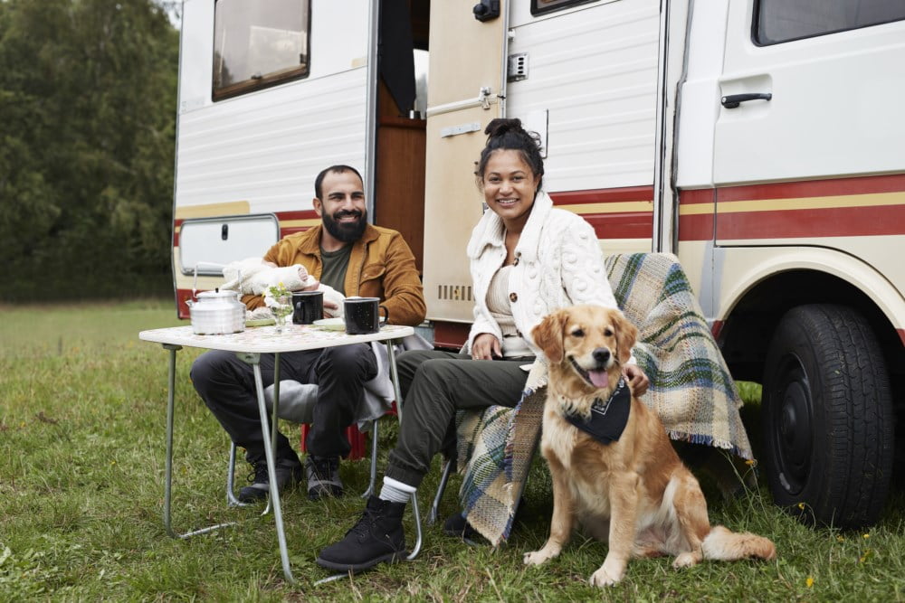 Eit par smiler og drikk kaffi ved eit bord utanfor bobilen sin med ein glad hund ved sidan av dei, omgitt av natur.
